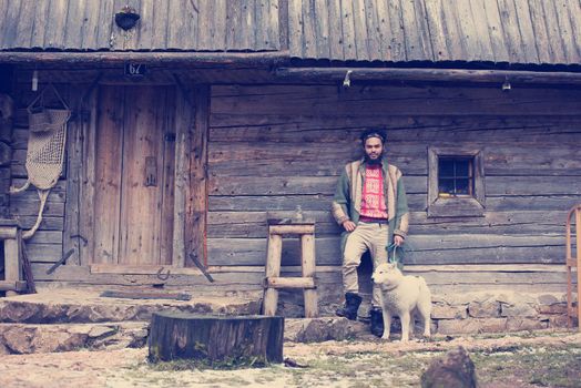 handsome young hipster man standing together with white husky dog in front of old vintage retro wooden house