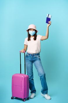 Full length of happy asian tourist, girl travelling abroad, posing with her suitcase in medical face mask, showing passport and tickets, blue background.