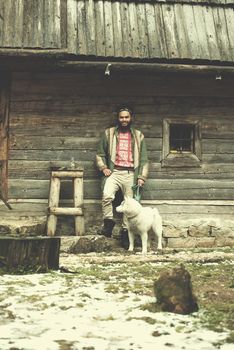 handsome young hipster man standing together with white husky dog in front of old vintage retro wooden house