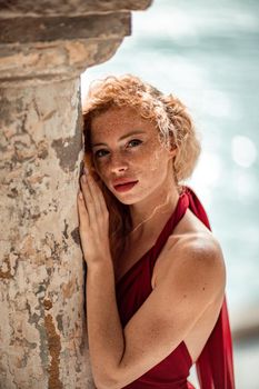 Outdoor portrait of a young beautiful natural redhead girl with freckles, long curly hair, in a red dress, posing against the background of the sea