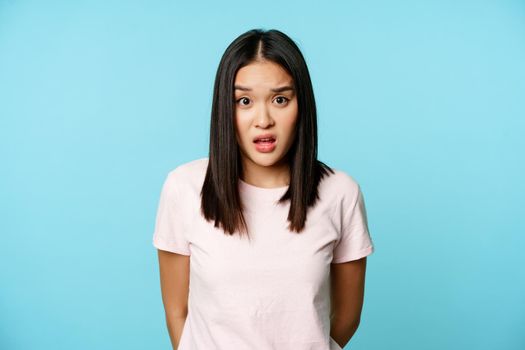 Image of asian korean girl look surprised, raising eyebrows confused, cant understand, standing over blue background.