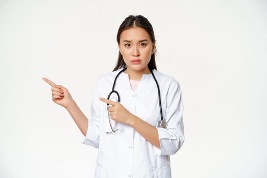 Sad and concerned female doctor, pointing fingers left, showing smth important, promo information, standing over white background.