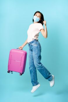 Full length portrait of happy asian woman tourist, jumping with suitcase, showing thumbs up, enjoying vacation going on tour, blue background.