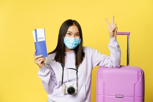 Japanese girl in medical mask, showing her passport and flight travel pass tickets, peace sign, posing with suitcase, going on vacation, yellow background.