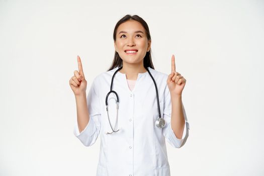 Happy smiling doctor, asian woman physician looking up with cheerful face expression, wearing medical uniform, white background.