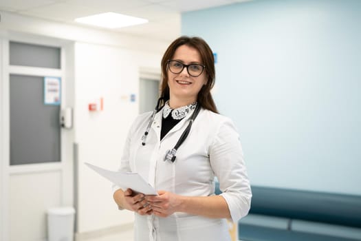 Female doctor standing in hospital corridor. Portrait of a young woman doctor in glasses and a white coat posing in a modern clinic. Proud professional woman doctor therapist looking at camera.