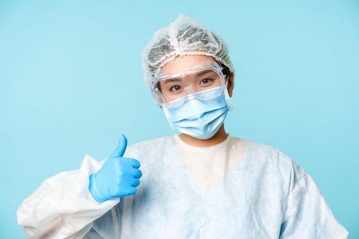 Smiling asian female nurse or doctor, wearing ppe, personal protective equipment, showing thumbs up, recommending vaccination, blue background.