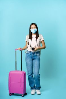 Happy asian girl traveller, tourist with suitcase and camera, going on vacation, wearing medical face mask during covid-19 restrictions, blue background.