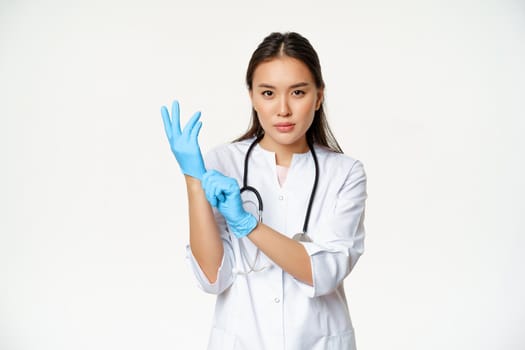 Confident female nurse, physician put on rubber medical gloves for patient clinical examination, standing serious in healthcare worker uniform, white background.
