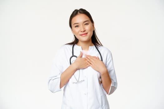 Image of female doctor caring for patients, holding hands on heart, smiling pleasant, standing in medical uniform against white background.