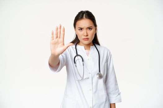 Asian doctor shows palm, stop gesture. Serious displeased female physician extend her arm, prohibit action, taboo sign, standing in uniform against white background.