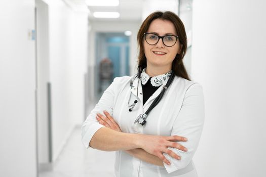 Female doctor standing in hospital corridor. Portrait of a young woman doctor in glasses and a white coat posing in a modern clinic. Proud professional woman doctor therapist looking at camera.