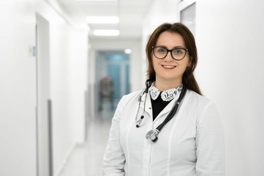 Confident smiling young woman doctor standing in medical institution. Proud professional doctor therapist woman in glasses looking at camera in hospital corridor. Portrait of general practitioner.