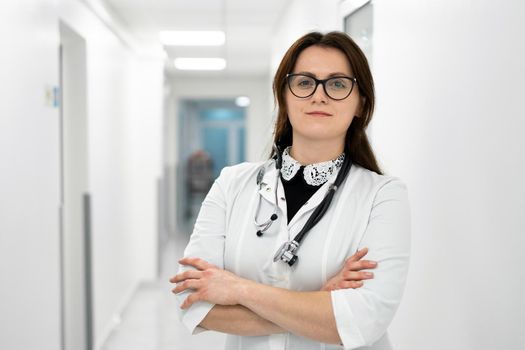 Headshot portrait of smiling millennial female doctor wearing medical uniform and stethoscope looking at camera in the corridor of a modern hospital. Healthcare concept, medical insurance.