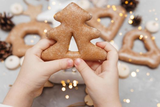 Children's hands hold a gingerbread in the shape of a Christmas tree. The concept of Christmas sweets, toys, garlands and bokeh. Decor for New Year holidays.