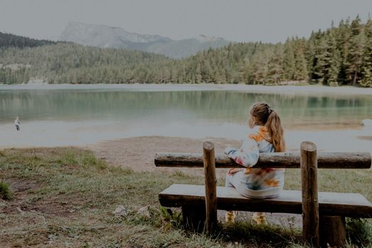 Young woman on shore of mountain lake. The back of girl against background of mountains. Young girl sitting near by beautiful Black Lake in Montenegro. Original wallpaper from summer morning.