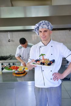 Handsome chef dressed in white uniform decorating pasta salad and seafood fish in modern kitchen