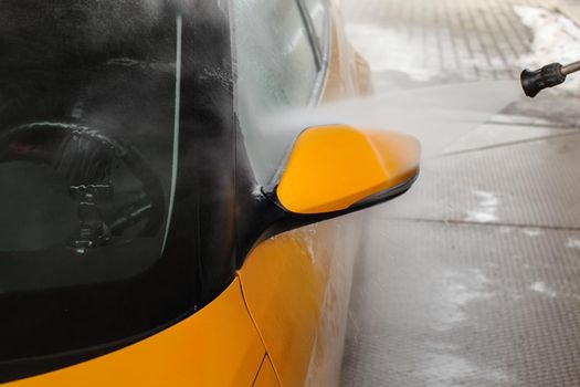 Yellow car high pressure water washing at carwash station. Jet water stream spraying on wing mirror.