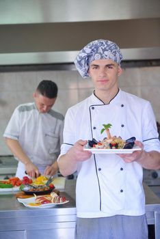 Handsome chef dressed in white uniform decorating pasta salad and seafood fish in modern kitchen