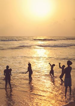 Kalutara, Sri Lanka - April 15, 2017: Silhouettes of people, mostly children playing on the beach in the sea in golden sunset evening light.