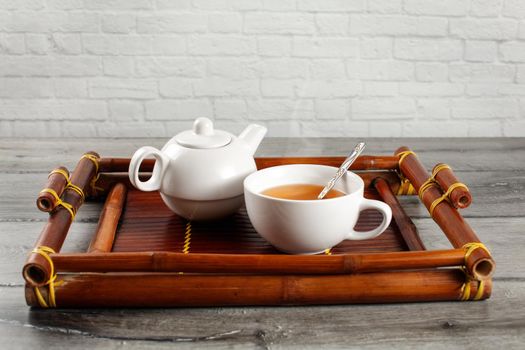 Small white ceramic teapot and cup of hot steaming black tea with silver spoon served on old bamboo tray. placed on gray wood desk.