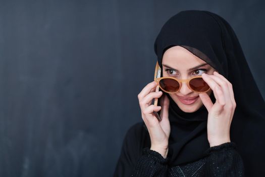 young modern muslim business woman using smartphone wearing sunglasses and hijab clothes in front of black chalkboard