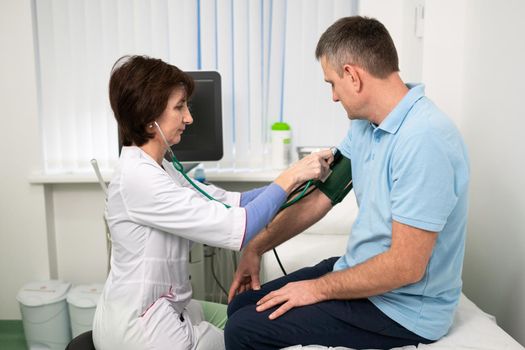 Female doctor measures blood pressure with a sphygmomanometer to male patient at medical examination in clinic office. Cardiologist checks blood pressure of male patient at cardiology hospital.