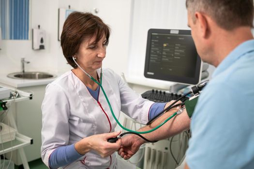 Female doctor measures blood pressure with a sphygmomanometer to male patient at medical examination in clinic office. Cardiologist checks blood pressure of male patient at cardiology hospital.