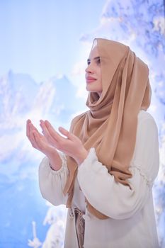 portrait of beautiful muslim woman in fashionable dress with hijab making traditional prayer to God, keeps hands in praying gesture isolated on modern winter mountains background representing modern islam fashion and ramadan kareem concept