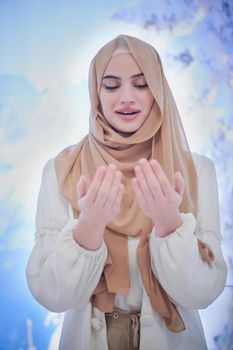 portrait of beautiful muslim woman in fashionable dress with hijab making traditional prayer to God, keeps hands in praying gesture isolated on modern winter mountains background representing modern islam fashion and ramadan kareem concept