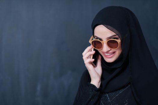 young modern muslim business woman using smartphone wearing sunglasses and hijab clothes in front of black chalkboard