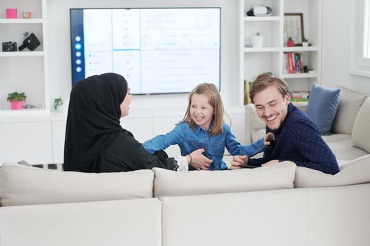 Happy Muslim family with daughter woman in traditional fashionable dress having fun and good time together while sitting on sofa during the month of Ramadan at modern home