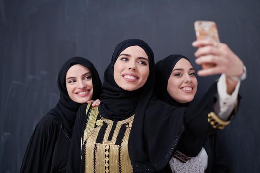 group of young beautiful muslim women in fashionable dress with hijab using mobile phone while taking selfie picture in front of black chalkboard representing modern islam fashion technology and ramadan kareem concept