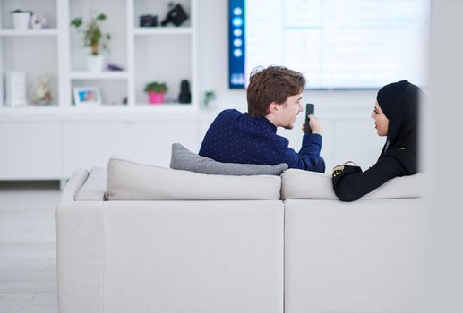 young muslim couple woman wearing islamic hijab clothes sitting on sofa watching TV together during the month of Ramadan at modern home