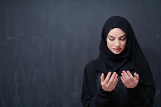 portrait of beautiful muslim woman in fashionable dress with hijab making traditional prayer to God keeps hands in praying gesture isolated on black chalkboard background representing modern islam fashion and ramadan kareem concept