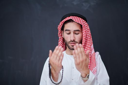 young arabian man in traditional clothes making traditional prayer to God, keeps hands in praying gesture in front of black chalkboard representing modern islam fashion and ramadan kareem concept