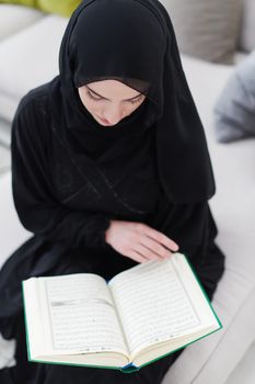 young traditional muslim woman reading Quran on the sofa before iftar dinner during a ramadan feast at home