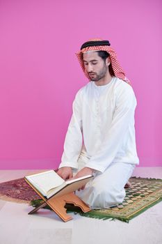 young arabian muslim man in traditional clothes reading holy book Quran on the praying carpet in front of pink wall before iftar dinner during a ramadan feast at home