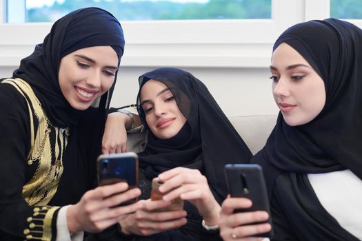 group of young beautiful muslim women in fashionable dress with hijab using mobile phone while sitting on the sofa near window at home representing modern islam fashion technology and ramadan kareem concept