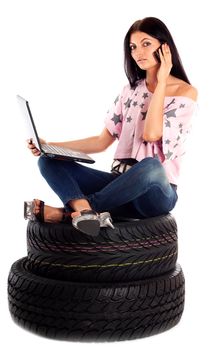 Young sexy woman with laptop and mobile sitting on the car wheels, isolated on white.