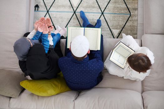 Traditional muslim family parents with children reading Quran and praying together on the sofa before iftar dinner during a ramadan feast at home top view