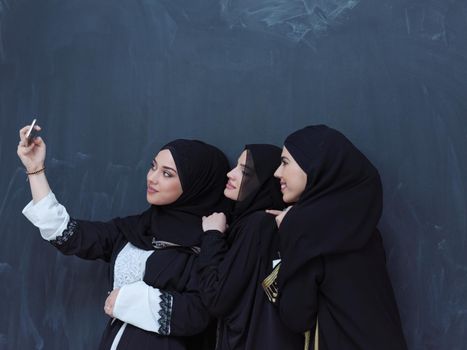 group of young beautiful muslim women in fashionable dress with hijab using mobile phone while taking selfie picture in front of black chalkboard representing modern islam fashion technology and ramadan kareem concept