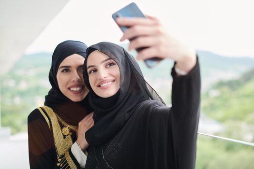 young beautiful muslim women in fashionable dress with hijab using mobile phone while taking selfie picture on the balcony representing modern islam fashion technology and ramadan kareem concept