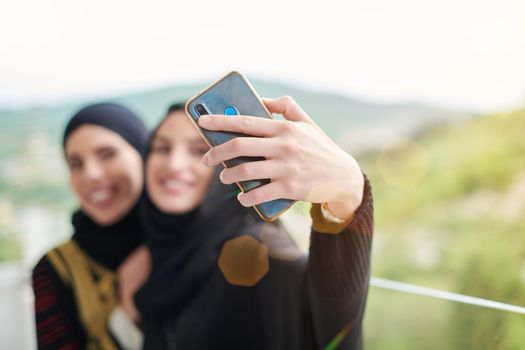 young beautiful muslim women in fashionable dress with hijab using mobile phone while taking selfie picture on the balcony representing modern islam fashion technology and ramadan kareem concept