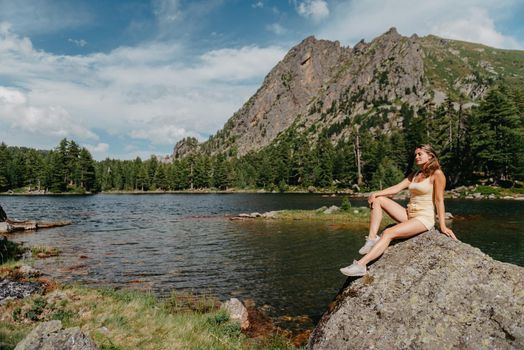 Tourist girl enjoys the magical view of the lake, coniferous forest and magical view sitting on big stone on the shore of a turquoise lake in the mountains. Hiking in the Natural Park. Black lake.