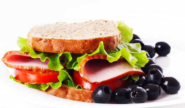 Big healthy sandwich with wholewheat bread, ham, tomatoes, olive and curly lettuce on the plate.