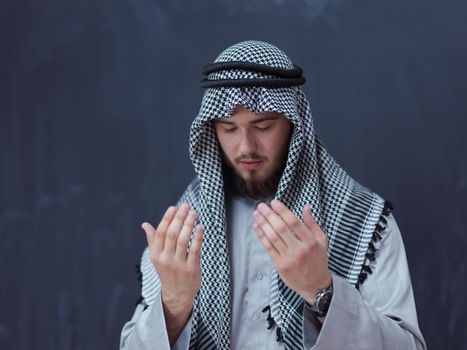 young arabian man in traditional clothes making traditional prayer to God, keeps hands in praying gesture in front of black chalkboard representing modern islam fashion and ramadan kareem concept