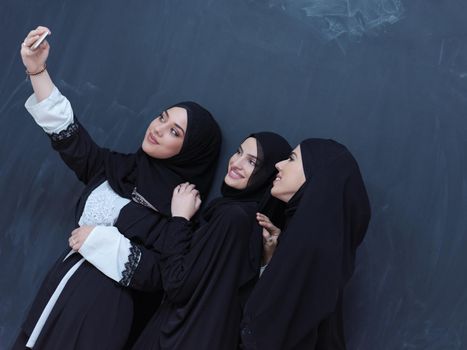 group of young beautiful muslim women in fashionable dress with hijab using mobile phone while taking selfie picture in front of black chalkboard representing modern islam fashion technology and ramadan kareem concept