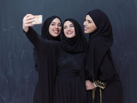 group of young beautiful muslim women in fashionable dress with hijab using mobile phone while taking selfie picture in front of black chalkboard representing modern islam fashion technology and ramadan kareem concept