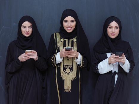 group of beautiful muslim women in fashionable dress with hijab using mobile phone in front of black chalkboard representing modern islam fashion technology and ramadan kareem concept